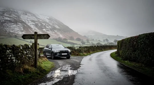 A rural British road splitting into two paths, one climbing into a snow-dusted highland track and the other continuing on a smooth wet tarmac lane, symbolising the choice between four-wheel drive and front-wheel drive.