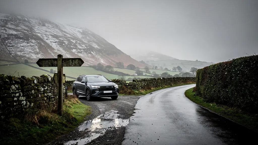 A rural British road splitting into two paths, one climbing into a snow-dusted highland track and the other continuing on a smooth wet tarmac lane, symbolising the choice between four-wheel drive and front-wheel drive.