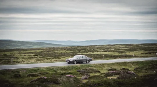 A single car driving along a winding road through vast, empty moorland under a dramatic sky, symbolizing a long solitary road trip.