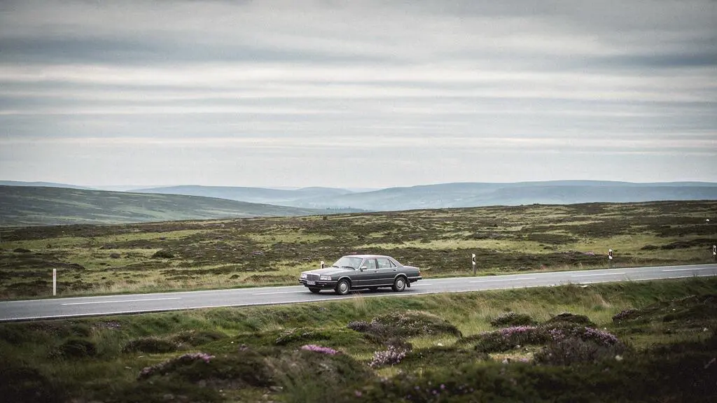 A single car driving along a winding road through vast, empty moorland under a dramatic sky, symbolizing a long solitary road trip.