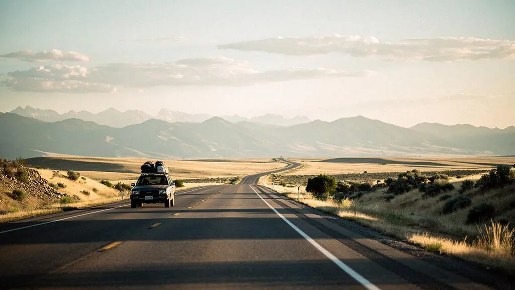 A car driving on a long open highway toward distant mountains at golden hour, symbolizing a three-week road expedition
