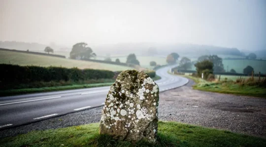 A moss-covered ancient stone marker standing quietly beside a deserted rural road at dawn, symbolizing hidden history just off the beaten path