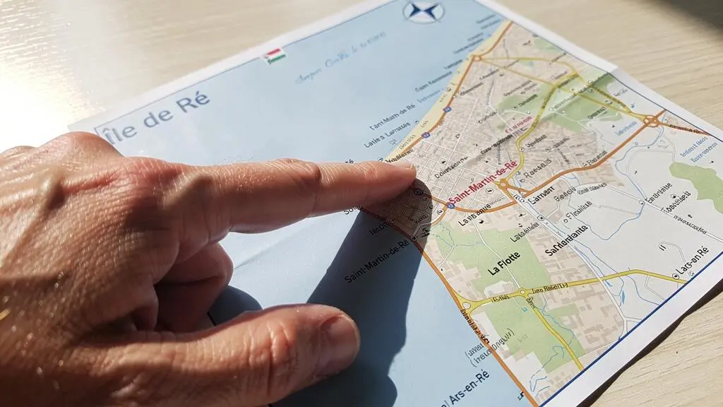 Close-up of a finger pointing at a specific location on a printed Île de Ré map spread on a wooden table, shallow depth of field with map text softly blurred