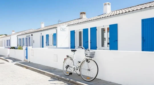 Wide coastal view of a contemporary white villa with blue shutters on Île de Ré, bicycle leaning against a stone wall in the foreground under bright summer sunlight
