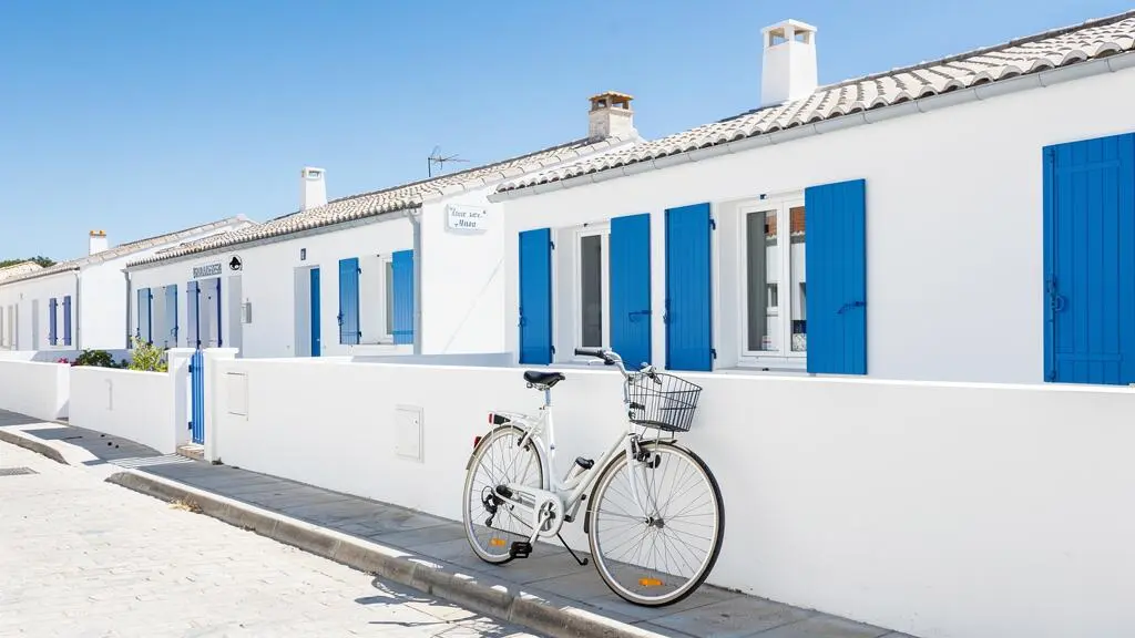 Wide coastal view of a contemporary white villa with blue shutters on Île de Ré, bicycle leaning against a stone wall in the foreground under bright summer sunlight